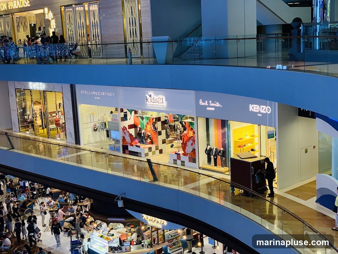 An interior view of a multi-level shopping mall. On the upper level, several storefronts are visible, including Gucci, Stella McCartney Kids, Kids21, Paul Smith Junior, and Kenzo Kids. Shoppers are seen browsing inside and outside these stores. Below, on a lower level, a busy food court area is visible with many people seated at tables and ordering food from various stalls. The mall features modern architecture with glass railings and escalators.