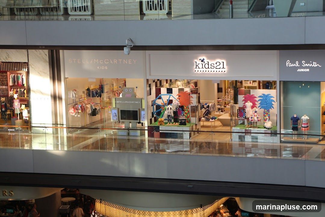 A view of multiple storefronts in a mall, featuring children's clothing. The stores visible are STELLAMcCARTNEY KIDS, kids21, and Paul Smith JUNIOR. Each store has displays of clothing and mannequins in their windows.