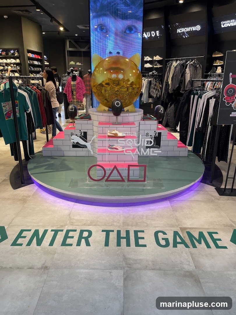 A retail store display featuring a large golden piggy bank filled with money, inspired by the Netflix series Squid Game. The display includes shoes, clothing racks, and the iconic Squid Game shapes (circle, triangle, square). The floor has green lettering that reads "ENTER THE GAME".
