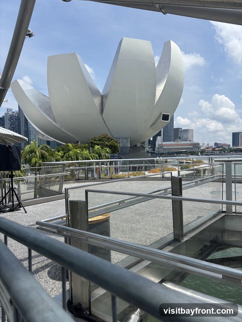 A wide shot of the ArtScience Museum in Singapore, with its distinctive lotus-shaped roof. The museum is surrounded by water and palm trees, with the city skyline in the background. The image is taken from a walkway with metal railings in the foreground.