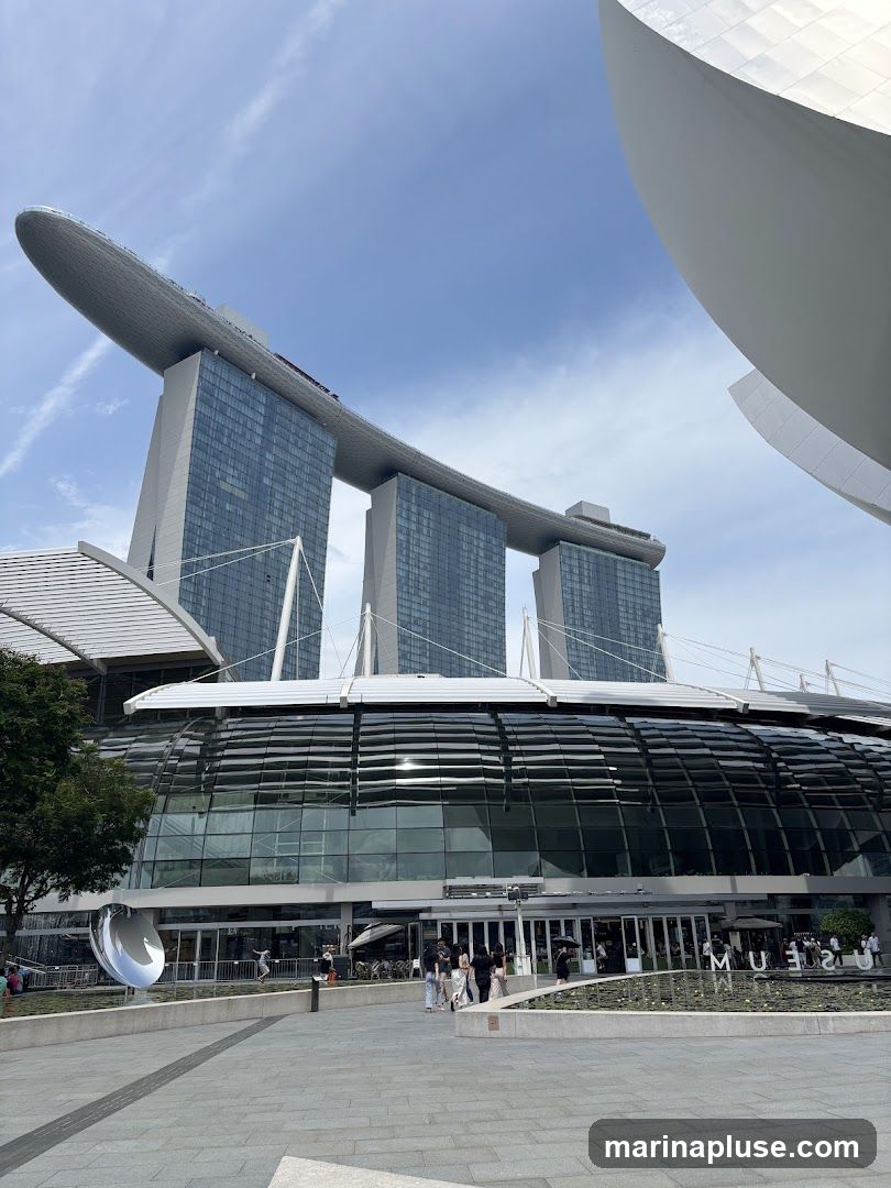 A wide shot of the Marina Bay Sands complex in Singapore, focusing on the museum building. The iconic three towers of the hotel rise in the background, connected by the SkyPark. The museum features a distinctive lotus-shaped glass dome. People are seen walking around the plaza in front of the museum.