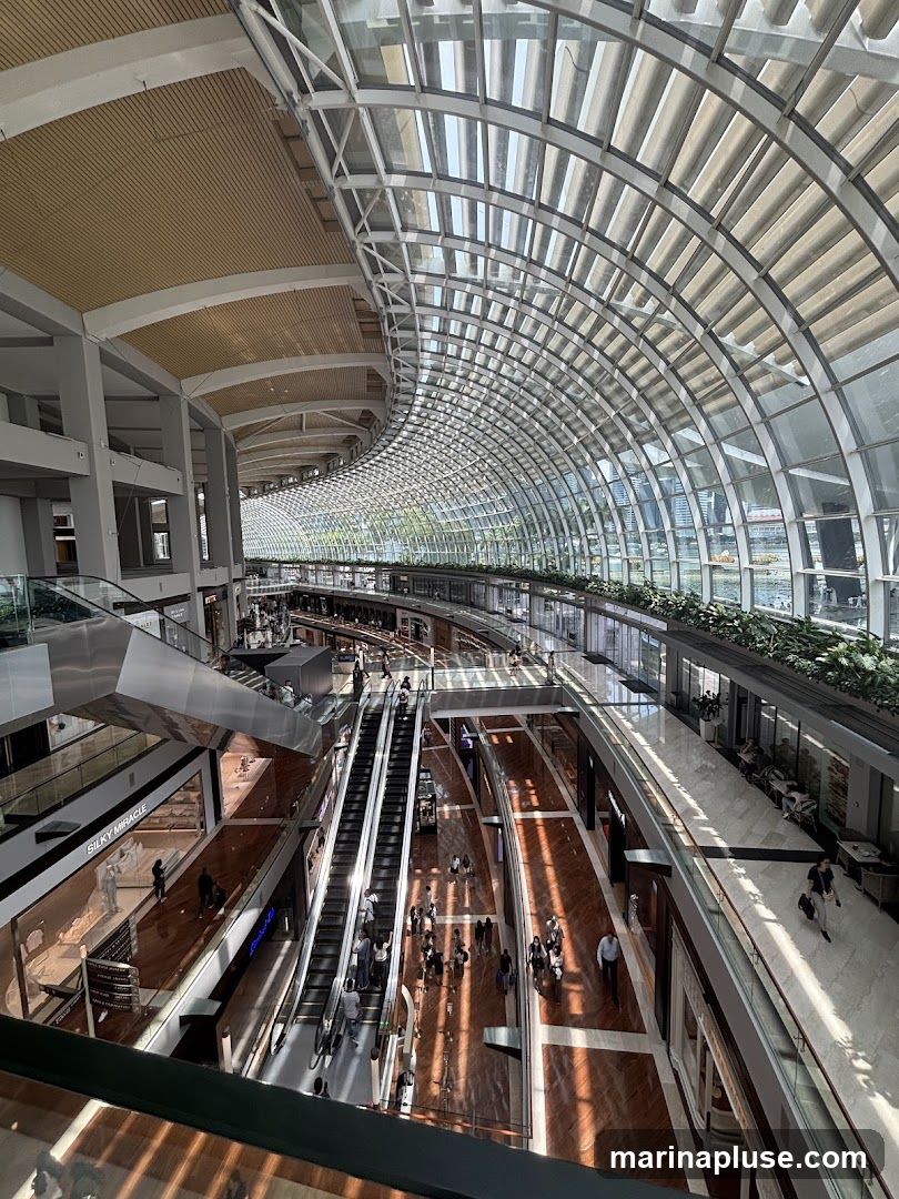 A wide-angle view of the interior of a large, modern shopping mall. The architecture features a curved glass roof that allows natural light to flood the space. Multiple levels of shops and escalators are visible, with shoppers moving between floors. The floors are made of polished wood, reflecting the sunlight streaming in. Greenery is incorporated along the upper levels, adding a touch of nature to the urban environment.