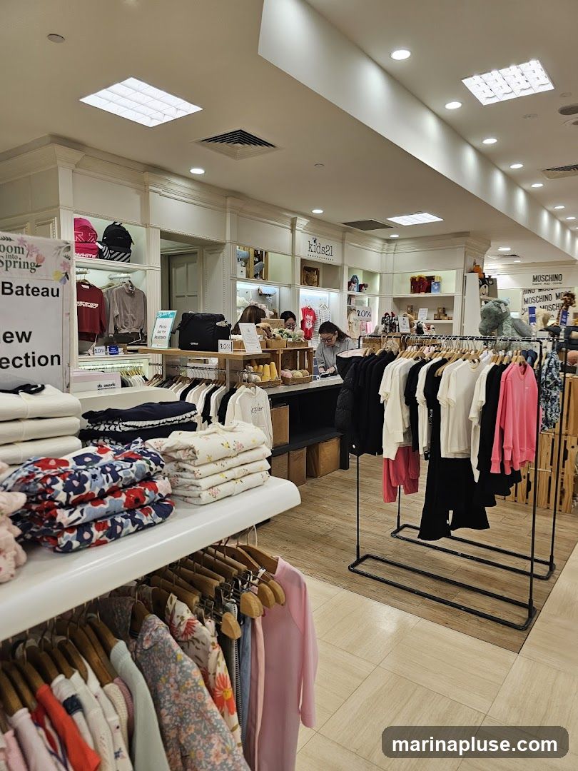 The image shows the interior of a children's clothing store. Racks of colorful clothes, including dresses, shirts, and sweaters, are displayed. Stacks of folded clothes are visible on shelves. Signage for "kids21" and "MOSCHINO" is present. Some items are marked with "20%" discounts.