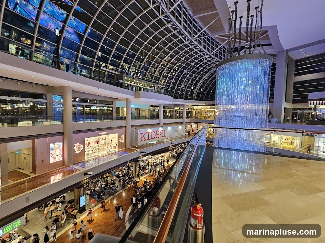 The image shows the interior of a large, multi-level shopping mall with a distinctive glass ceiling that reflects colorful lights. A large, illuminated crystal chandelier hangs from the ceiling, casting a blue glow. Several shops are visible on different levels, including one with a sign that reads "KIDS21" and another with "MONNALISA". People are seen walking and dining in the mall. The overall ambiance is bright and bustling.