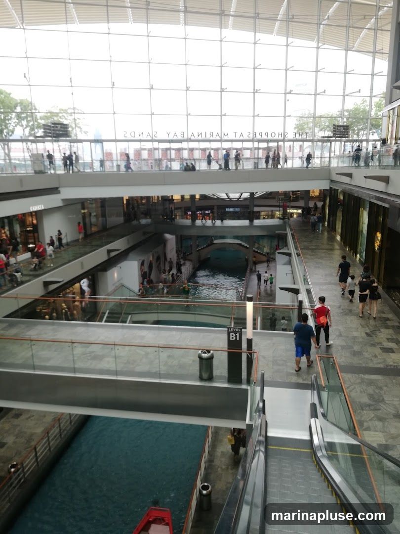 An interior view of a multi-level shopping mall with a canal running through it. People are seen walking on various levels and on escalators. The mall has a modern design with glass railings and a large glass ceiling. The text "THE SHOPPES MARINA BAY SANDS" is visible on the upper level.