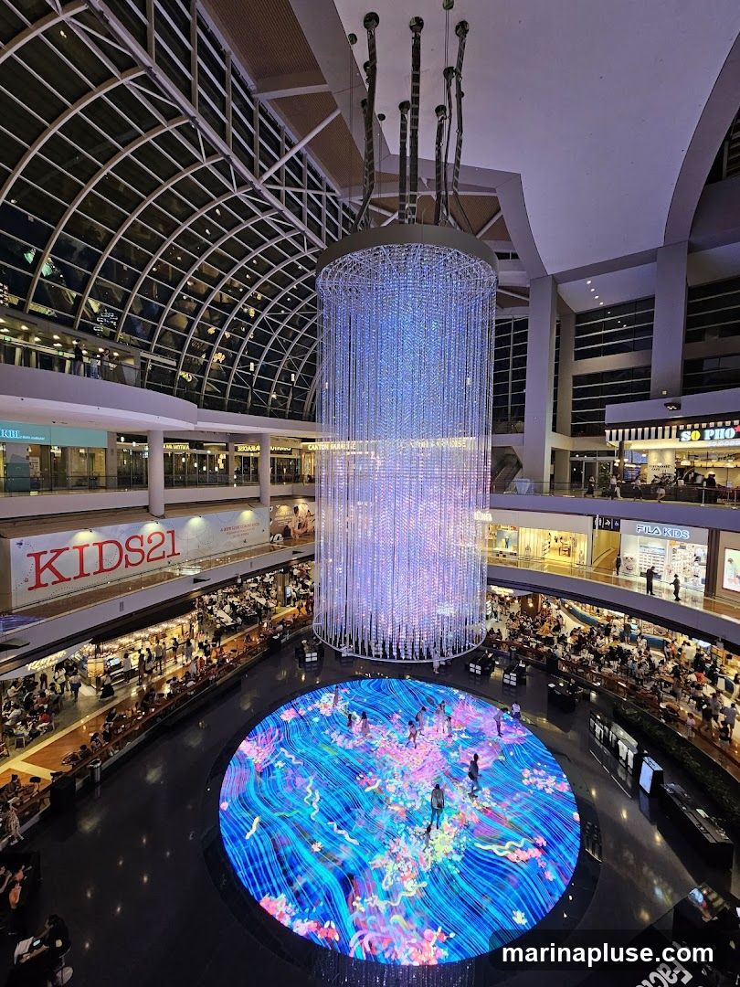 An interior view of a large, modern shopping mall featuring a massive, illuminated chandelier hanging from the ceiling. Below the chandelier is a circular digital display on the floor, showing a vibrant, abstract pattern with people walking on it. The mall has multiple levels with shops and restaurants visible, and many people are browsing or dining. The architecture includes a large, arched glass roof.