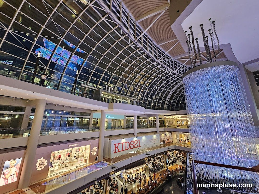 A wide-angle view of the interior of a multi-level shopping mall, featuring a large, illuminated crystal chandelier, a glass-paneled ceiling with a digital display, and various shops and dining areas. The architecture is contemporary, with clean lines and a spacious feel. People are seen browsing shops and dining at tables below.