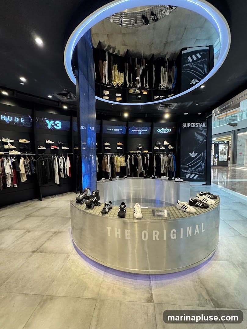 The interior of an Adidas store featuring a circular display in the center with the text "THE ORIGINAL" and various Adidas shoes. The walls display clothing racks and shelves with more Adidas merchandise, including shoes and apparel. Brands like "Y-3" and "JOHN ELLIOTT" are visible on the wall displays. The ceiling has a reflective surface showing a display of clothing.