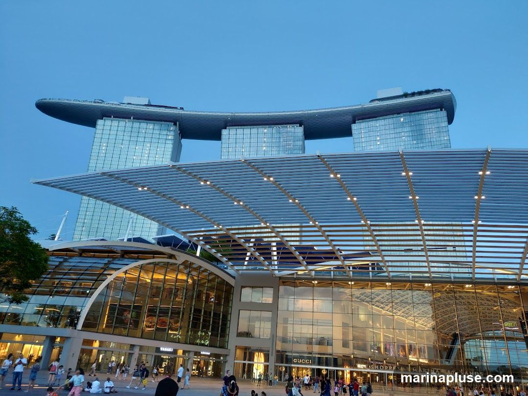 A wide shot of the Marina Bay Sands complex in Singapore at dusk. The iconic three towers topped with a ship-like structure are visible in the background. In the foreground, the shopping mall's facade with its glass and metal architecture is illuminated. People are seen walking and sitting in the plaza area. Signs for various shops, including Gucci and The Shoppes Marina Bay Sands, are visible.