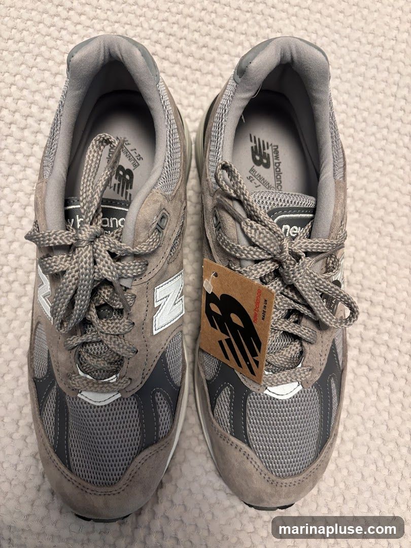 A pair of grey New Balance sneakers with white laces and a brown tag attached to the right shoe. The tag has the New Balance logo and text that reads "New Balance MADE IN UK". The shoes appear to be new and unworn.