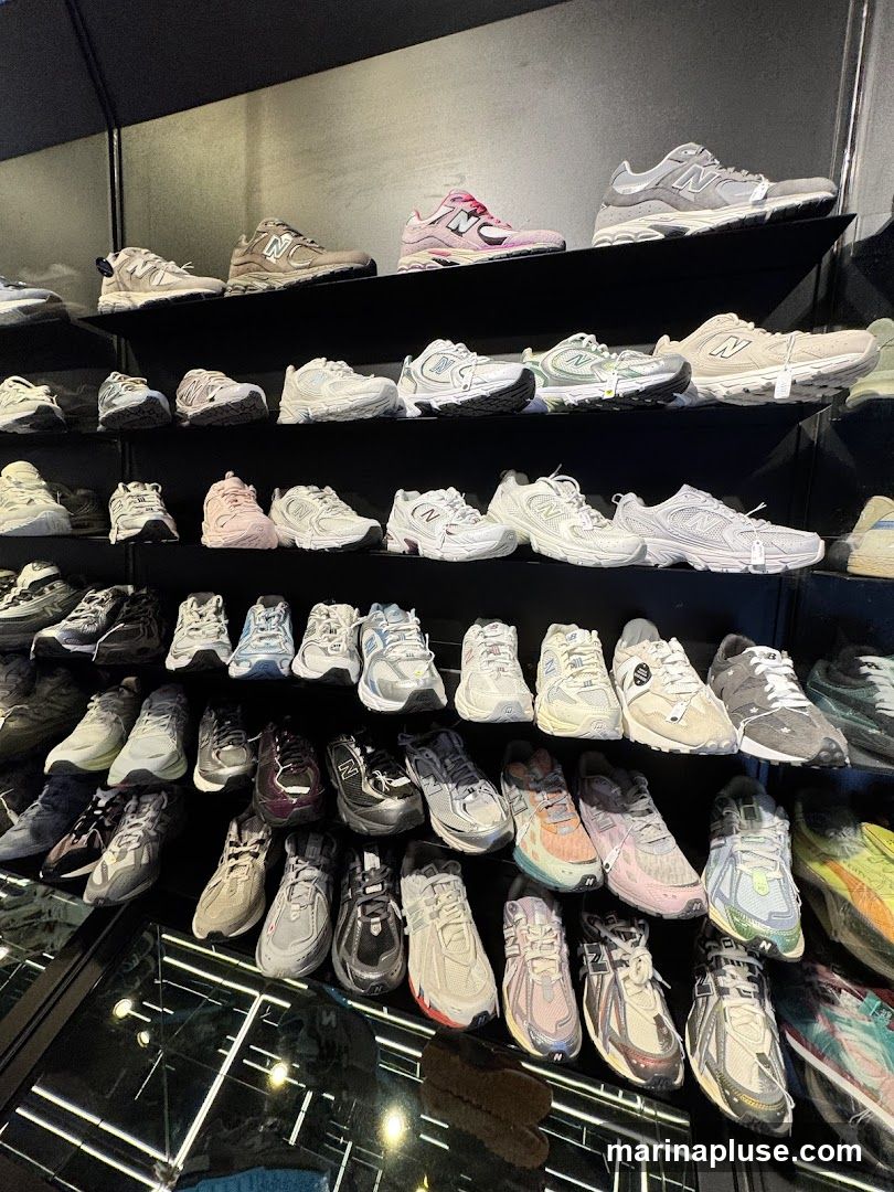 A wall display of numerous New Balance sneakers on black shelves. The shoes are arranged in rows, showcasing a variety of styles and colors, including grey, white, pink, and beige. Some shoes have tags attached. The lighting is dim, highlighting the footwear.