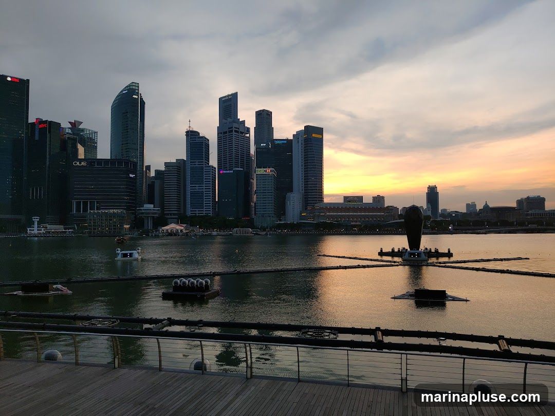 A wide shot of a city skyline at dusk, with tall buildings reflected in the water. The sky is a mix of grey clouds and warm orange and yellow hues from the setting sun. In the foreground, a wooden boardwalk with a metal railing overlooks the water. Several floating structures are visible in the water, including one that resembles a large, dark, abstract sculpture with radiating arms. There are also smaller floating platforms and what appears to be a swan-shaped boat. The overall scene captures a serene urban landscape as day transitions into night.