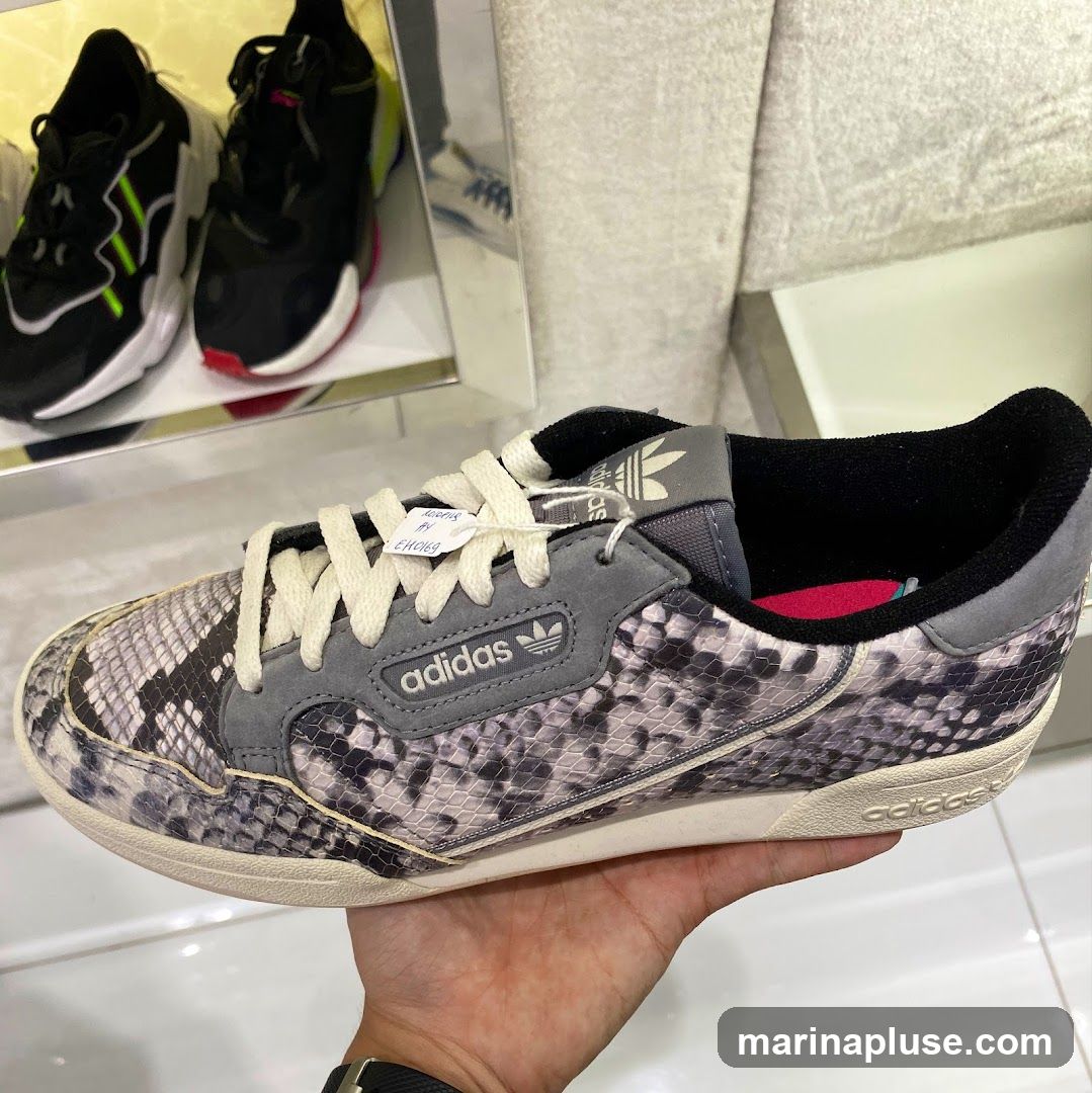 A hand holds a grey Adidas sneaker with a snakeskin pattern. The sneaker has white laces and a white sole. In the background, two other pairs of Adidas sneakers are visible on a shelf.