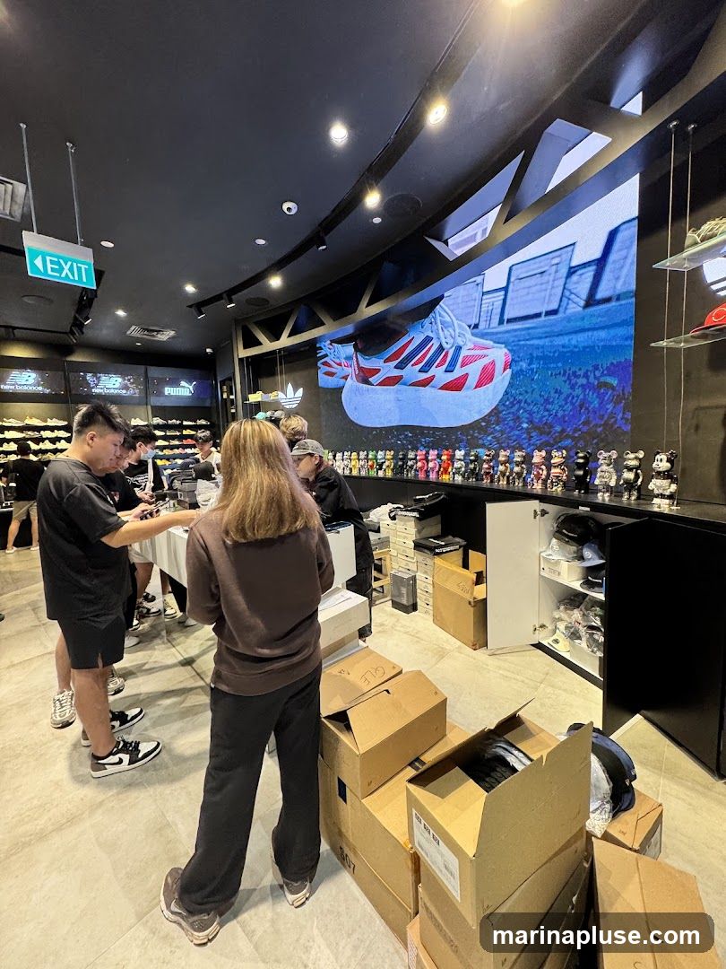 The interior of a modern shoe store with several customers and staff. A large screen displays an advertisement for a sneaker. Shelves on the left display various shoe brands like New Balance and Puma. The floor is tiled, and the ceiling is black with track lighting. Cardboard boxes are stacked in the foreground, suggesting recent deliveries or inventory.