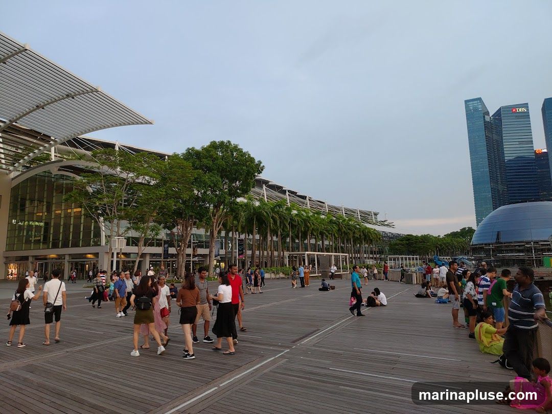 A wide shot of a bustling waterfront promenade with many people walking and sitting. In the background, a modern building with a glass facade is visible, lined with palm trees. Further in the distance, several skyscrapers stand tall against a cloudy sky. The promenade is made of wooden planks, and there are various groups of people enjoying the outdoor space.