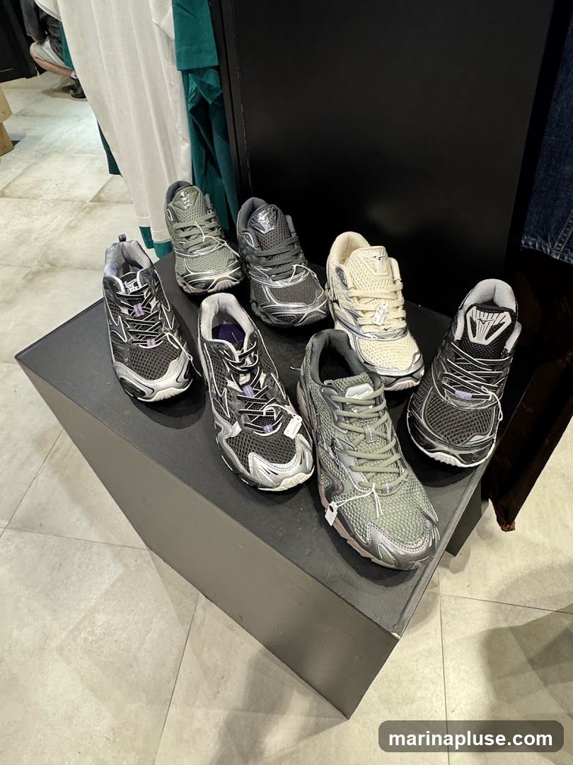 A display of several pairs of athletic shoes in various colors, including grey, black, and cream, are arranged on a dark, angled pedestal. The shoes appear to be running or training sneakers with mesh uppers and contrasting sole details. Some shoes have small white tags attached. In the background, clothing items hang on racks, suggesting an indoor retail environment.