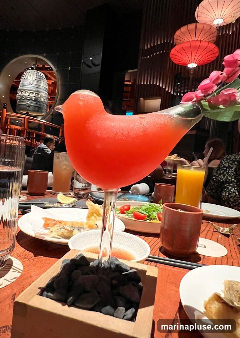 A close-up shot of a vibrant red, frozen cocktail served in a unique bird-shaped glass. The drink is garnished with a pink flower and green leaves. In the background, a table is set with various dishes, including tempura, a salad with cherry tomatoes and greens, and what appears to be dumplings. The restaurant's interior features traditional Japanese-inspired decor, with lanterns hanging from the ceiling and a large decorative bell. The overall ambiance suggests an upscale dining experience.