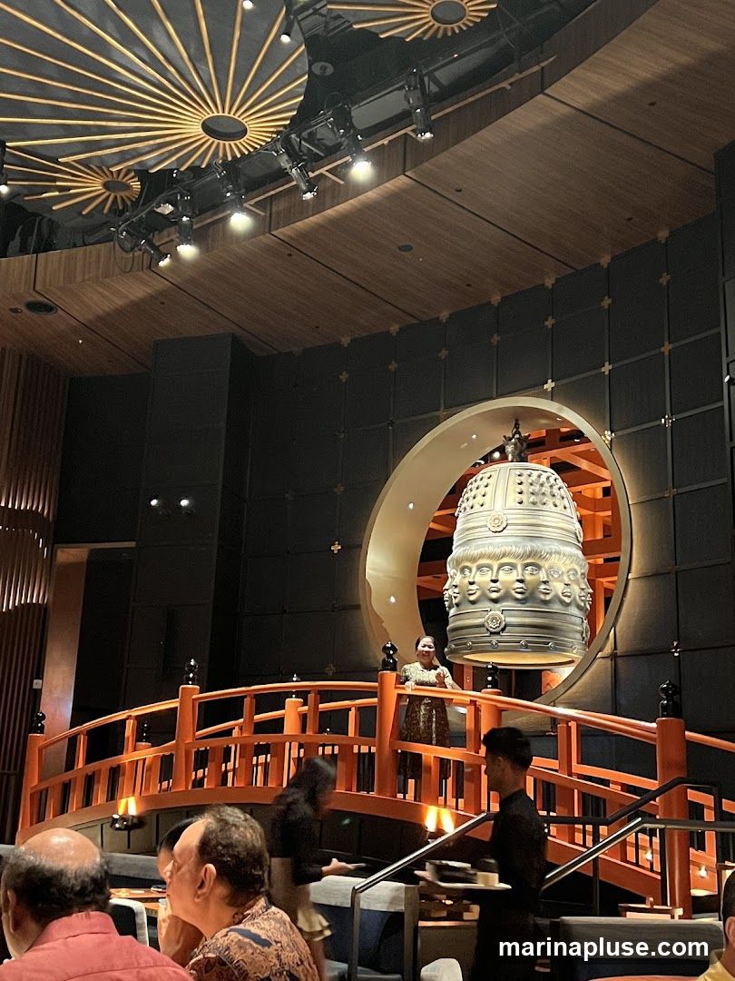 The image shows the interior of a restaurant with a distinctive Asian-inspired design. A large, ornate bell with multiple faces is a central decorative element, suspended within a circular archway. A wooden bridge with orange railings spans across the dining area, with people walking on it and seated at tables below. The ceiling features unique sunburst-like light fixtures and wooden paneling. Staff members are visible, one carrying a tray. The overall ambiance appears sophisticated and atmospheric.
