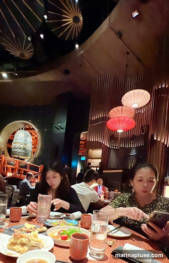 A dimly lit restaurant interior with patrons seated at tables. The decor features decorative ceiling lights resembling sunbursts and hanging paper lanterns in red and white. A large, ornate bell is visible in the background. People are dining, with plates of food and drinks on the tables.