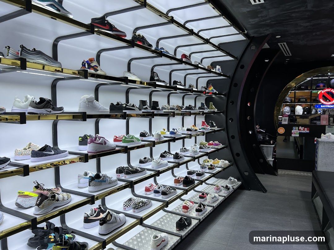 A well-lit shoe store interior with shelves displaying a wide variety of sneakers. The shelves are arranged in a curved, modern design, with white backlighting highlighting the footwear. Different brands and styles of athletic shoes are visible, from high-tops to low-tops, in various colors and designs. In the background, a circular archway leads to another section of the store, where more merchandise and a counter are visible. Signage for "BAYFRONT" and "UNDERGROUND" can be seen in the background.