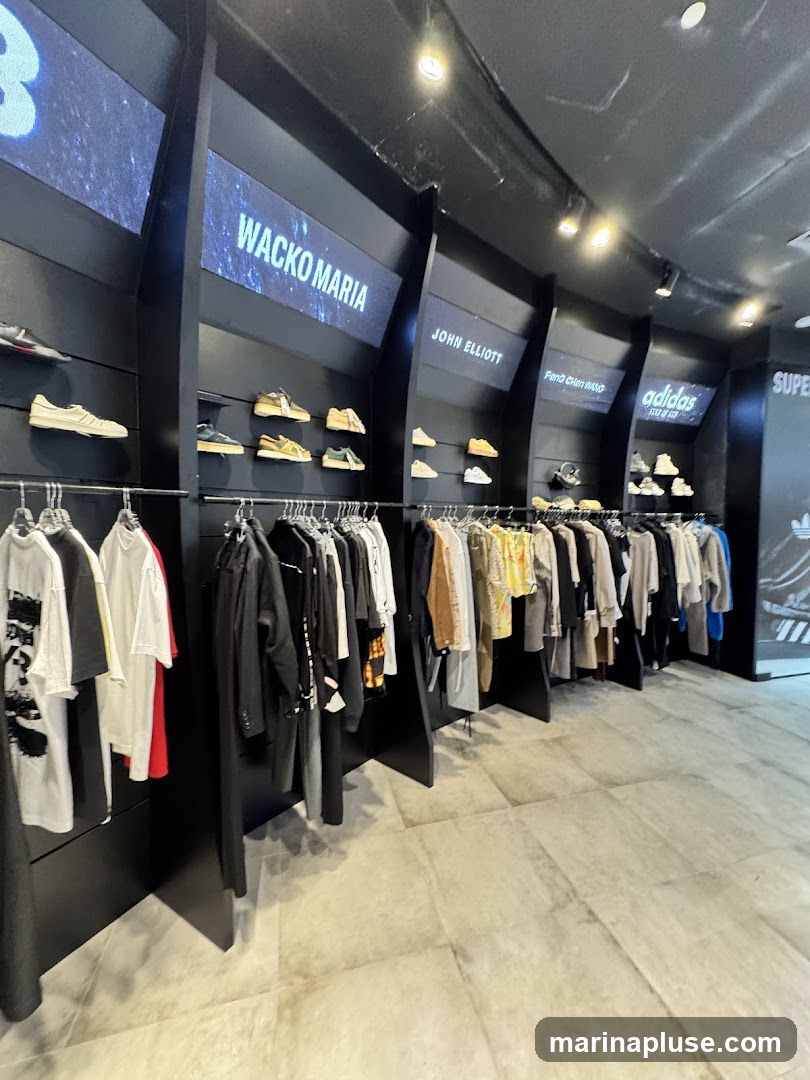 A well-lit clothing store interior featuring a display of apparel and footwear. The walls are black, with illuminated panels displaying brand names such as "WACKO MARIA", "JOHN ELLIOTT", "Feng Chen Wang", and "adidas". Shelves above the clothing racks showcase various sneakers. The clothing racks display a selection of t-shirts, jackets, and pants. The floor is tiled with large, light-colored tiles.