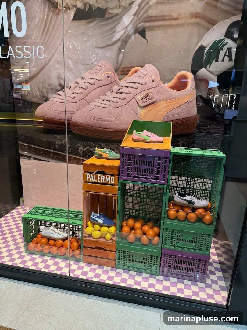 A store window display featuring Puma Palermo sneakers. The display includes several pairs of sneakers in various colors, arranged on top of and inside colorful crates. Oranges and lemons are also used as props in the display. In the background, a large image of a pair of pink Puma sneakers and a soccer ball are visible. Text on the window reads "10 CLASSIC" and "PALERMO" on one of the crates.