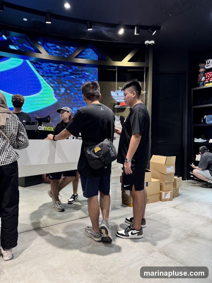 Several customers are standing at a counter in a brightly lit retail store. A large digital screen displays a blue and green abstract pattern behind the counter. There are shelves with merchandise and boxes stacked in the background. One person is wearing a hijab.