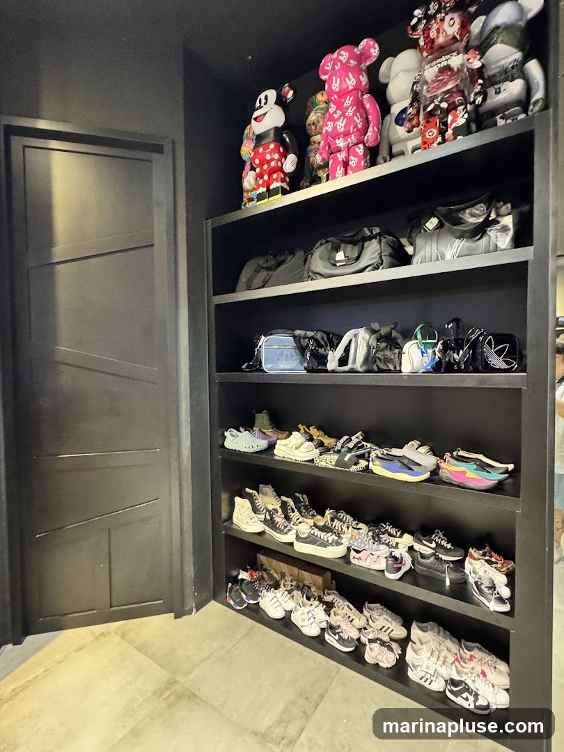A tall, black shelving unit displays various items. The top shelf holds several large collectible figures, including a Mickey Mouse figure and pink and white bearbrick-style figures. The second shelf contains several black duffel bags and backpacks. The third shelf is filled with smaller bags and pouches, some with visible branding like Adidas. The bottom three shelves are dedicated to an extensive collection of sneakers and shoes, neatly arranged. To the left of the shelves is a dark, paneled door.