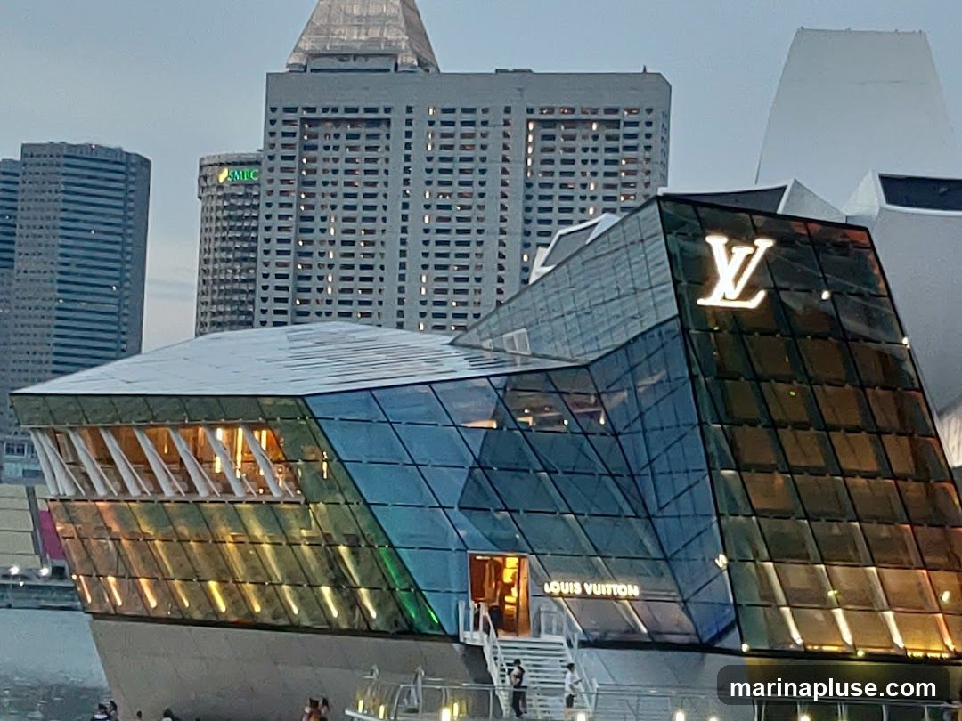 A modern, angular glass building with the Louis Vuitton logo prominently displayed on its facade. The building is situated by the water, with other city buildings in the background. The glass panels reflect the surrounding lights and sky, creating a colorful and dynamic exterior.
