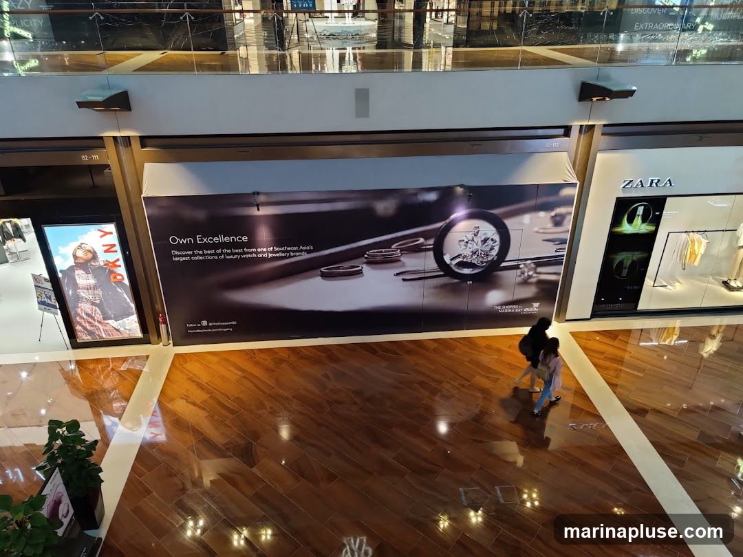 An overhead view of a shopping mall interior. On the left, a DKNY advertisement is displayed on a large screen. In the center, a large banner advertises luxury watches and jewelry with the text "Own Excellence." To the right, the ZARA store is visible with mannequins displaying clothing. Two people are walking across the polished wooden floor.