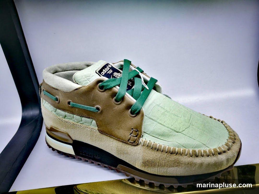 A close-up studio shot of a single Adidas shoe. The shoe has a light green textured toe box, brown leather accents, and a canvas-like material on the sides. It features green laces and a white sole with black and brown detailing. A label with "adidas" and "ZX 10000 C" is visible on the tongue.