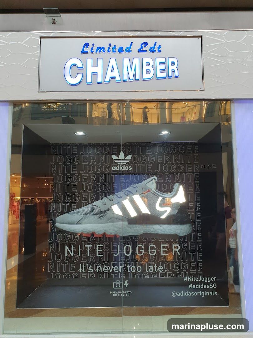 A store window display featuring a pair of Adidas Nite Jogger shoes. The display has the text "Limited Edt CHAMBER" above the window. The window itself is decorated with the repeating text "NITE JOGGER" and an image of the shoe. Below the shoe image, it says "It's never too late." and provides instructions to "TAKE A PHOTO WITH THE FLASH ON". Hashtags and social media handles for Adidas are also visible.
