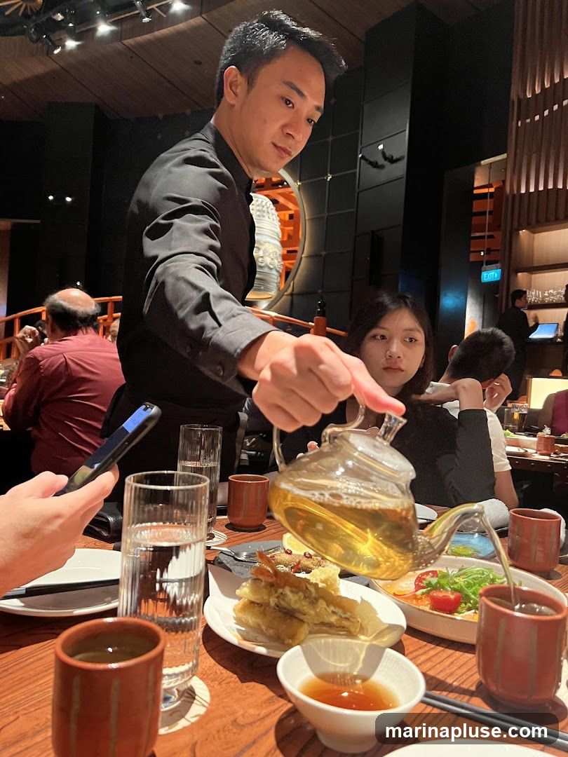 A waiter in a black uniform pours tea from a glass teapot into a ceramic cup at a table in a dimly lit restaurant. There are several plates of food on the table, including what appears to be tempura and a salad. Other diners are visible in the background.
