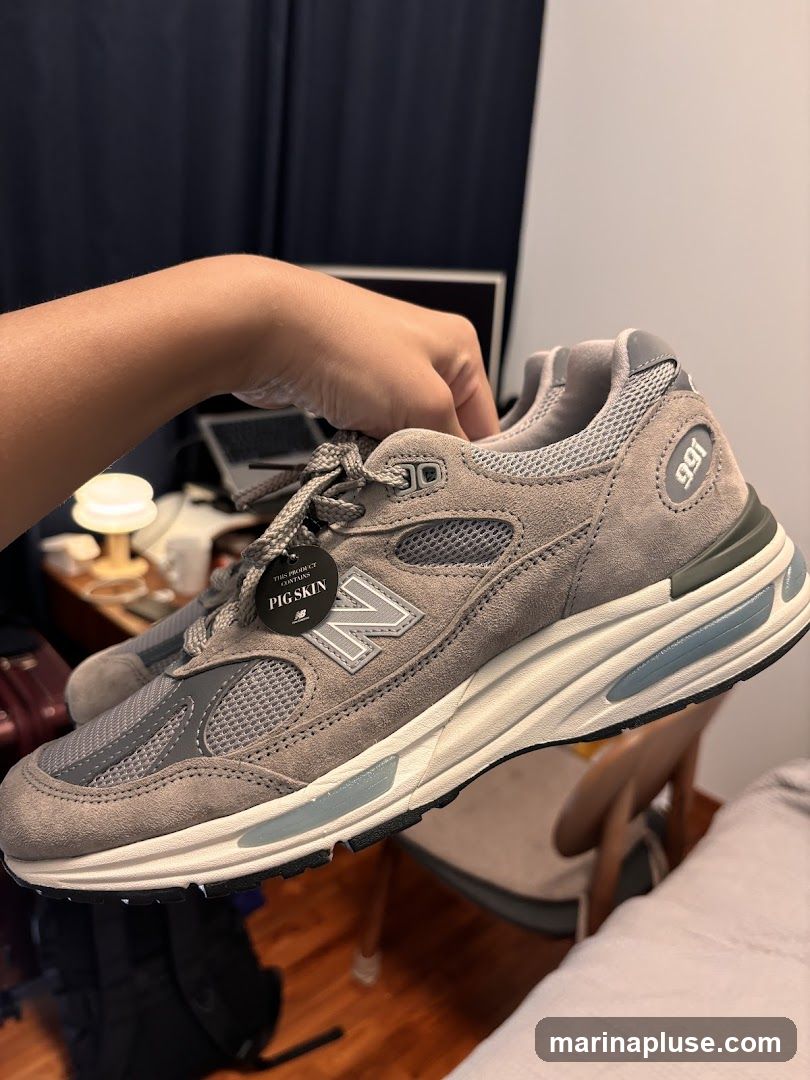 A person is holding a pair of grey New Balance 991 sneakers. The sneakers are made of suede and mesh with white soles and light blue accents. A tag on the shoe reads "THIS PRODUCT CONTAINS PIG SKIN" and displays the New Balance logo. The background shows a desk with a laptop, a lamp, and a chair.
