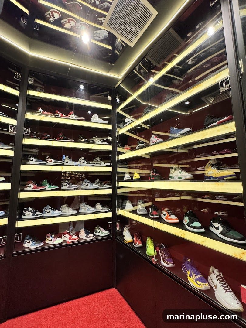 A well-lit display case filled with numerous pairs of sneakers, organized on shelves. The sneakers are of various brands and styles, with some appearing to be collectible or limited edition. The display case has mirrored surfaces and is illuminated by strip lighting, creating a modern and premium aesthetic. The floor is covered with a red carpet.