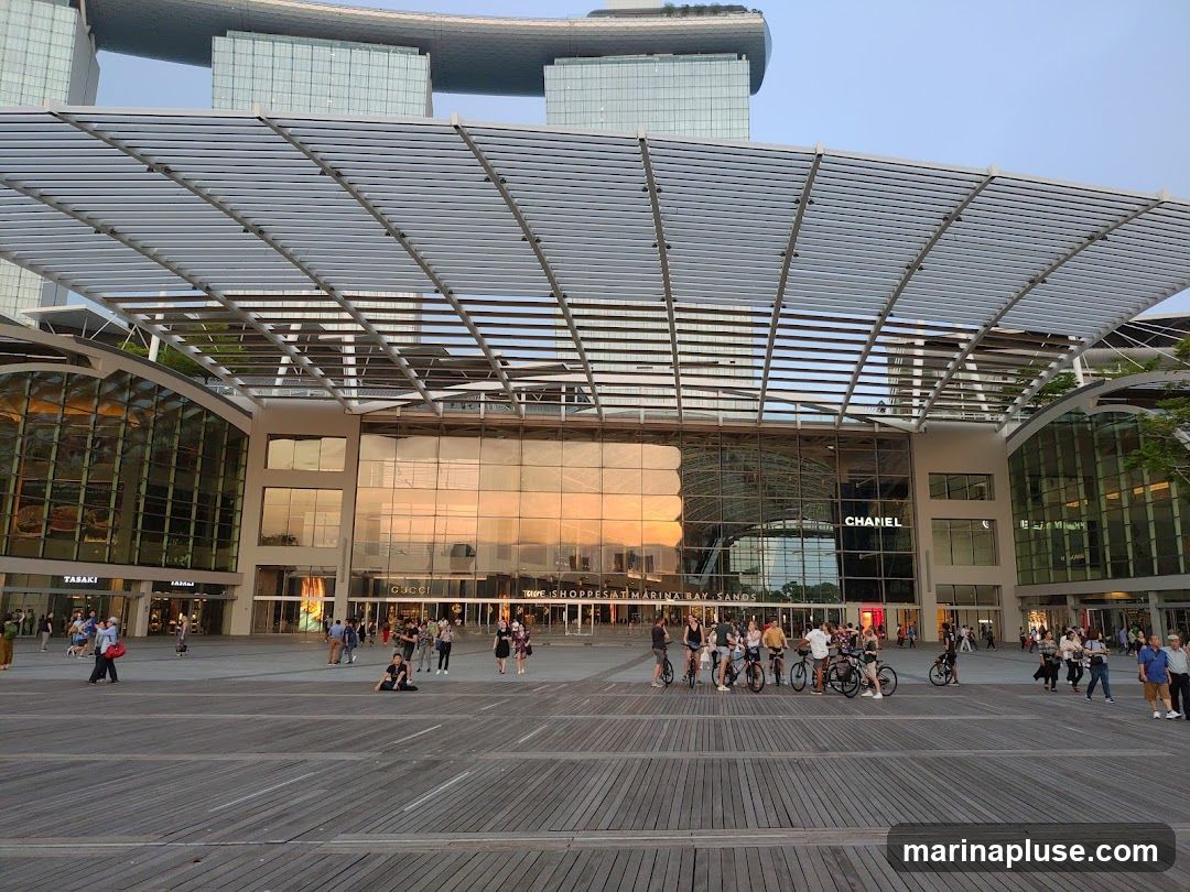 A wide shot of the exterior of The Shoppes at Marina Bay Sands, a luxury shopping mall. The building features a modern architectural design with a large glass facade that reflects the sunset. Several high-end brand names, including Chanel and Gucci, are visible on the storefronts. People are seen walking and cycling in the plaza in front of the mall. In the background, the iconic Marina Bay Sands hotel towers are visible.