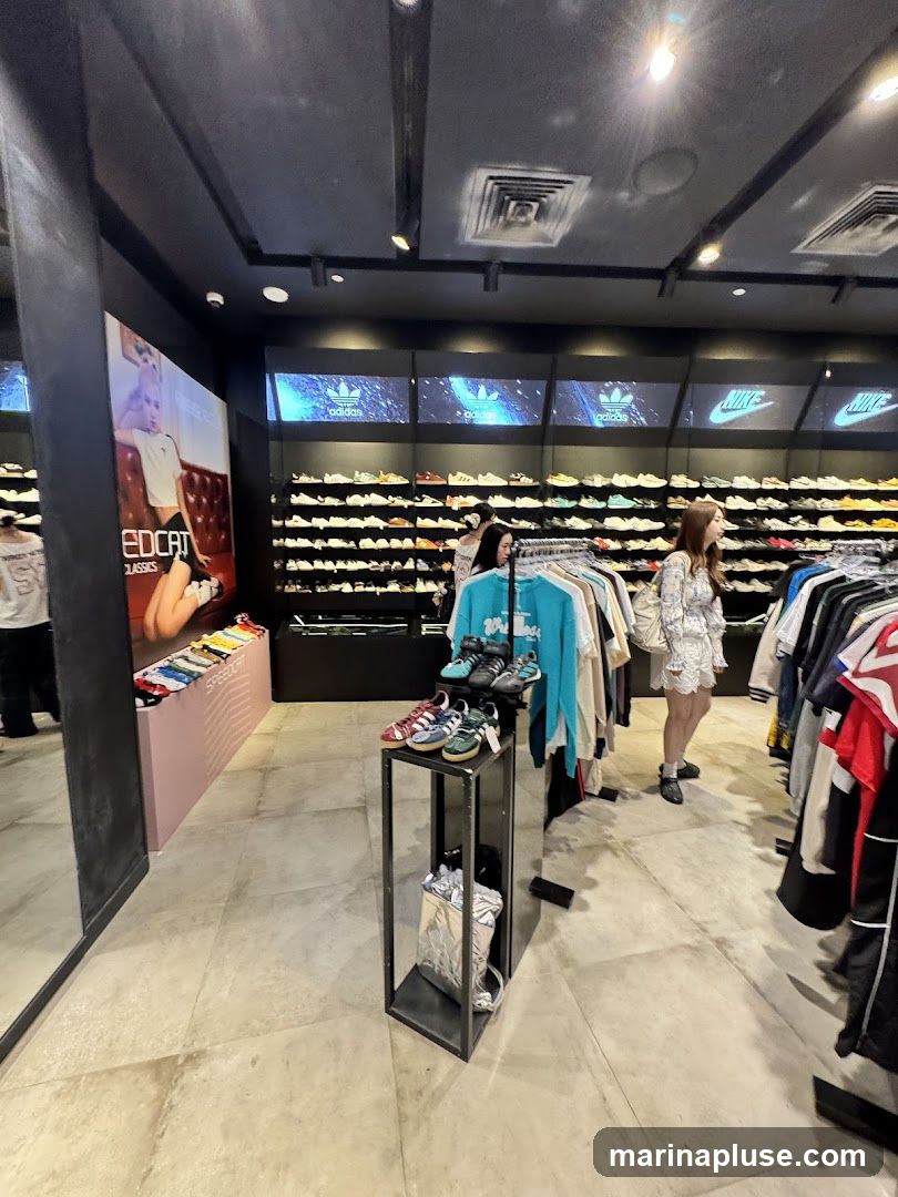 The interior of a brightly lit sneaker store. Racks of shoes are displayed on shelves along the walls, with prominent Adidas and Nike logos visible above. Several people are browsing the merchandise, which includes sneakers, t-shirts, and other apparel. A large poster featuring a model is on the left side of the image.
