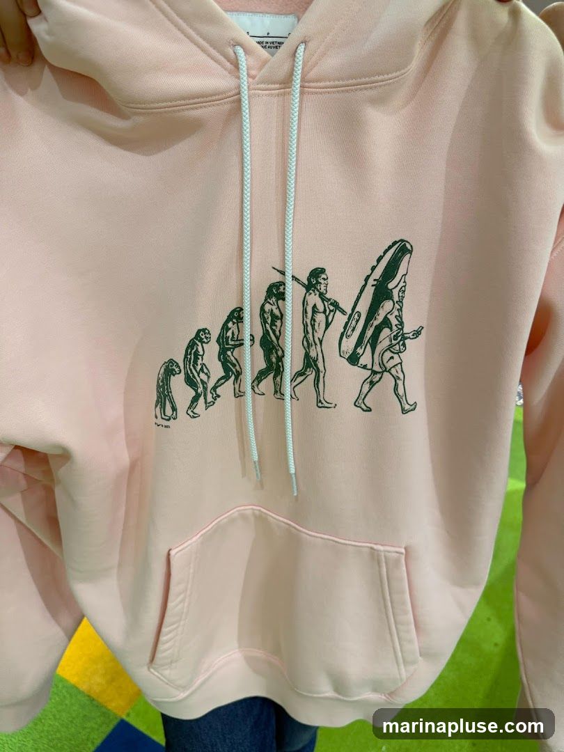 A light pink hoodie with a graphic depicting human evolution, culminating in a figure carrying a sneaker. The graphic is in dark green. The hoodie has a front pocket and white drawstrings.