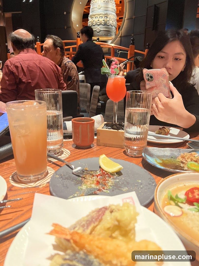 A group of people are seated at a table in a restaurant, with food and drinks on the table. In the background, a large decorative bell is visible, along with a server attending to other guests. The focus is on the dining experience, with close-ups of tempura, drinks, and plates.