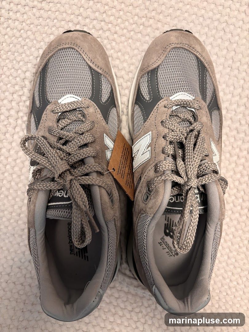 A top-down view of a pair of grey New Balance sneakers with white laces. The shoes are made of suede and mesh. A brown tag with text is attached to the laces of the left shoe. The New Balance logo is visible on the side of the shoes and on the insoles.