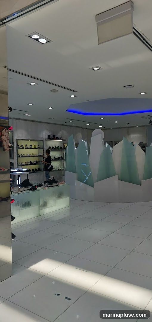 The interior of a modern shoe store with shelves displaying various footwear. A customer is browsing the merchandise. The store features a clean, minimalist design with white tiled floors, bright overhead lighting, and blue accent lighting on the ceiling. A large mirrored pillar is visible on the left side of the image. Decorative, angular partitions are placed in the center of the store.