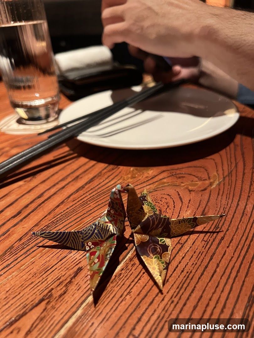 Two decorative origami cranes are placed on a wooden table. In the background, a glass of water, chopsticks, and a plate are visible, along with a person's hands.