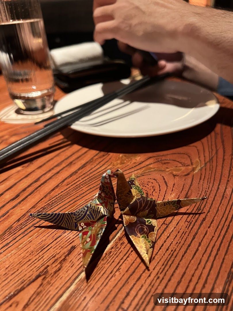 Two decorative origami cranes are placed on a wooden table. In the background, a glass of water, chopsticks, and a plate are visible, along with a person's hands.