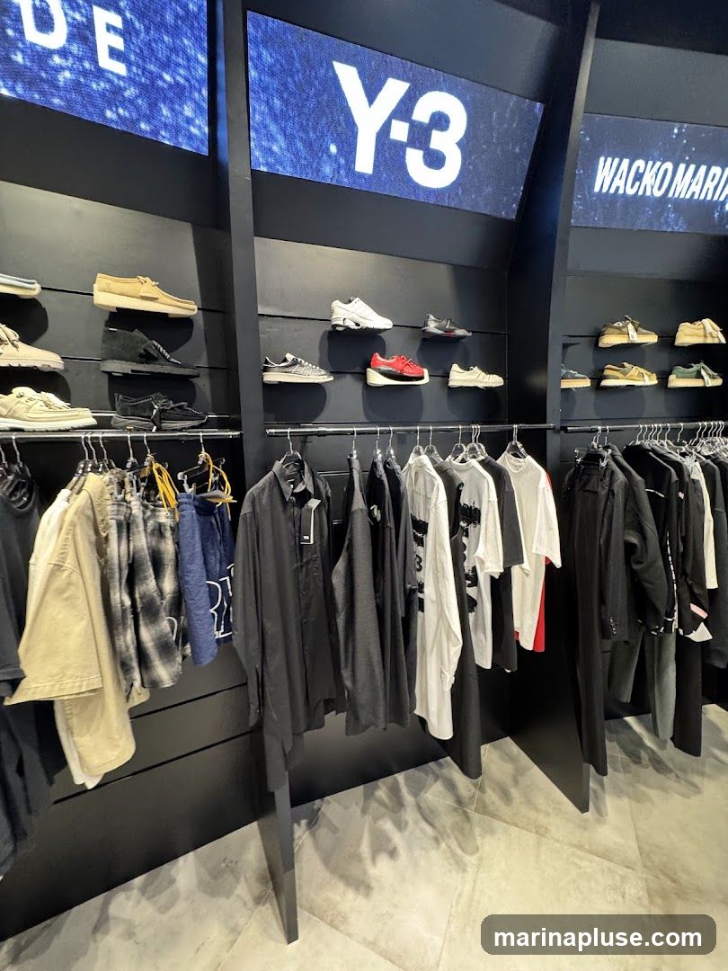 A retail store interior with clothing and shoes displayed on shelves and racks. Digital screens above display brand logos, including "Y-3" and "WACKO MARIA". Shoes are arranged on shelves, and various garments like shirts, jackets, and pants are hung on racks. The store has a modern, minimalist aesthetic with black walls and shelving, and a grey tiled floor.