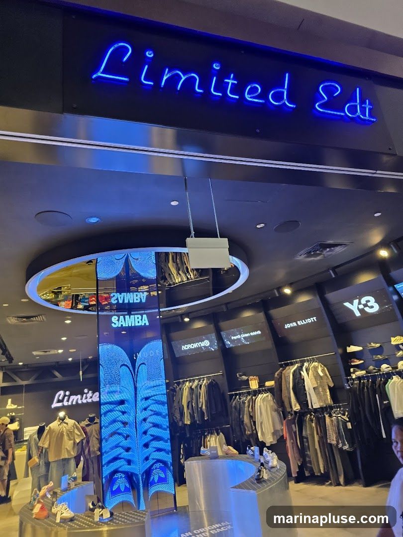 The image shows the exterior of a store with a neon blue sign that reads "Limited Edt". Inside, clothing racks display various apparel, and shoes are showcased on display stands. A large vertical screen shows an advertisement for Adidas Samba. Other brand names like Y-3, John Elliott, and Acronym are visible on the walls.