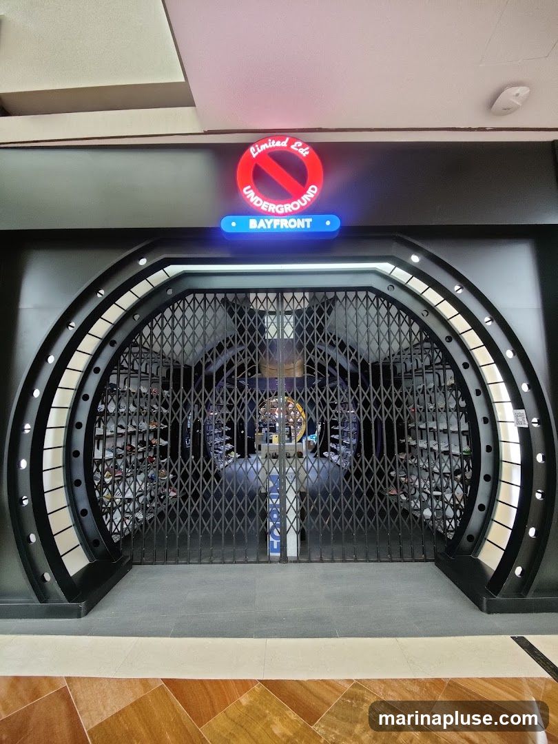 The exterior of a shop with a large arched entrance. The entrance is covered by a black metal security gate. Above the entrance, a neon sign reads "Limited Edt Underground" with a red circle and slash symbol, and below it, a blue illuminated sign reads "BAYFRONT". Through the gate, shelves stocked with shoes are visible.