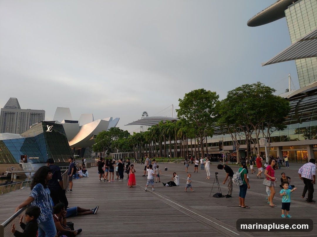 A crowd of people are seen on a wooden boardwalk in front of a modern building with a distinctive lotus-like roof structure. A Louis Vuitton store is visible on the left side of the building, with its logo displayed. The sky is overcast, and palm trees line the walkway. Several individuals and groups are standing, sitting, and walking, enjoying the outdoor space. In the background, other contemporary buildings and a Ferris wheel can be seen.
