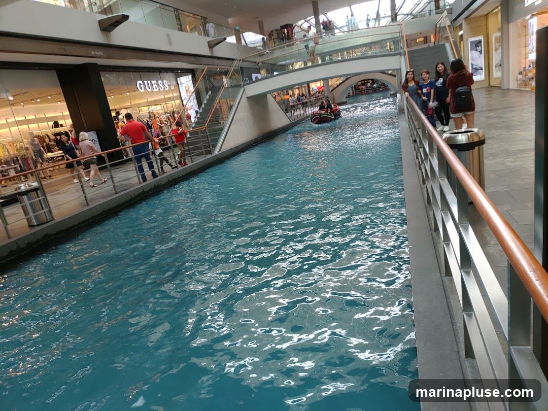 An indoor canal with bright blue water flows through a modern shopping mall. A small boat, resembling a gondola, navigates the waterway. People are visible on the walkways surrounding the canal, some looking at the shops, others observing the boat. A store with the prominent "GUESS" logo is visible on the left side of the image. The architecture suggests a large, upscale commercial space with multiple levels and bridges.