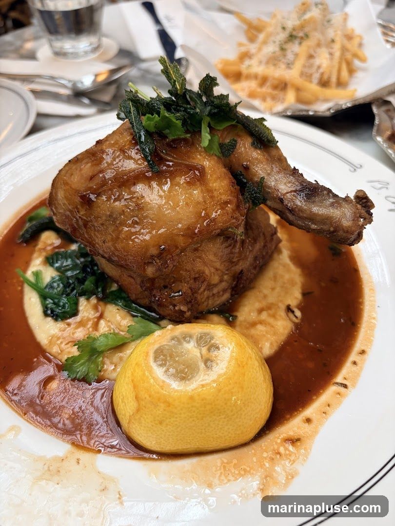 A close-up shot of a plate of food featuring a half roasted chicken served over creamy polenta. The chicken is glazed and garnished with fresh herbs. A halved lemon sits in the rich brown sauce surrounding the dish. In the background, a basket of french fries topped with cheese is visible, along with a glass of water.