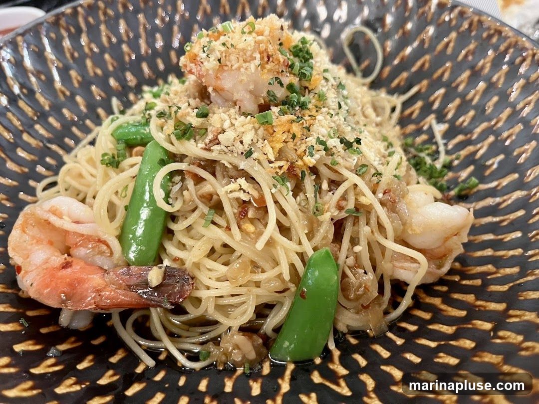 A close-up shot of a pasta dish with shrimp and snap peas, served in a decorative dark bowl. The pasta is thin and appears to be tossed in a light sauce, topped with breadcrumbs and chopped herbs. Several large shrimp are visible, along with bright green snap peas. The bowl has a textured, patterned design.